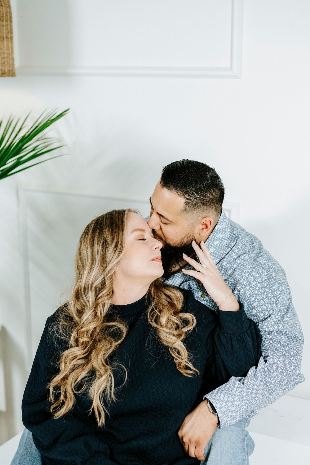 Intimate couple in casual wear sharing a tender moment on white couch with palm plant in background.