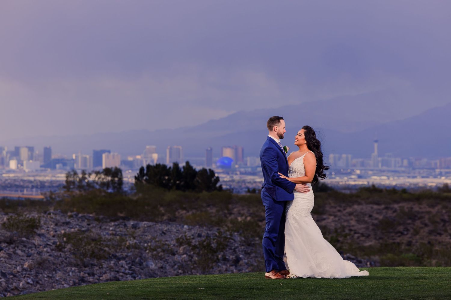A couple shares a romantic moment in front of a scenic desert landscape with rocky outcrops and lush greenery.