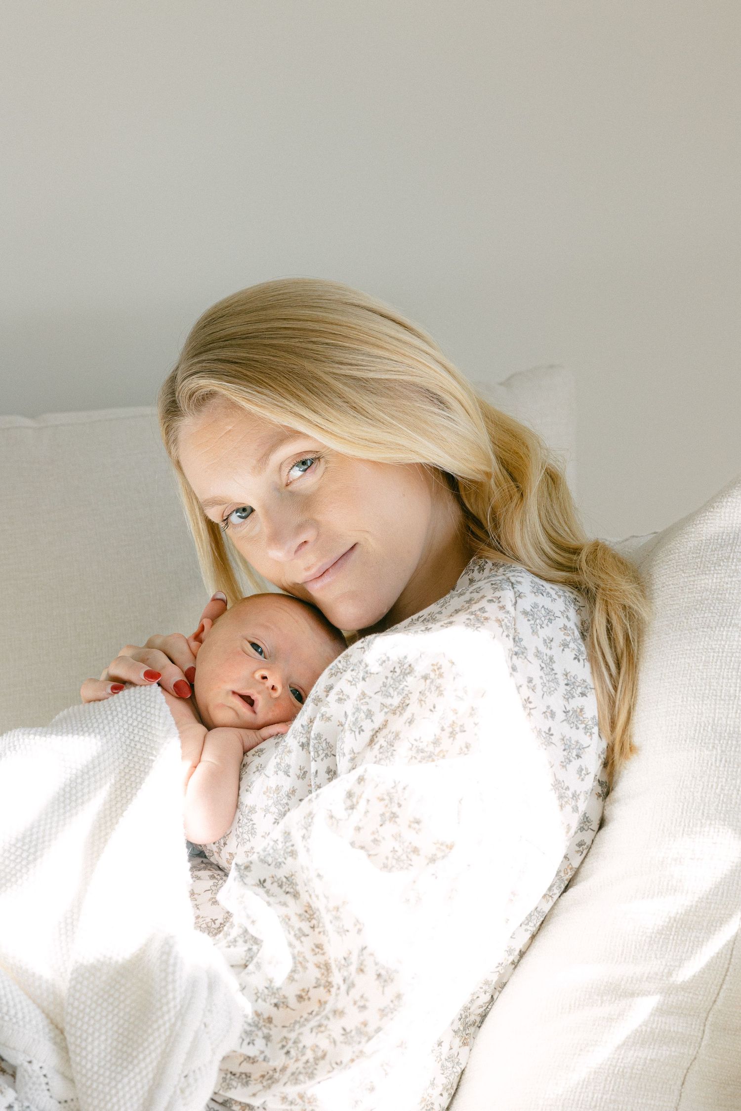 A tender moment shows a mother in white holding her sleeping newborn baby wearing a white bow headband.
