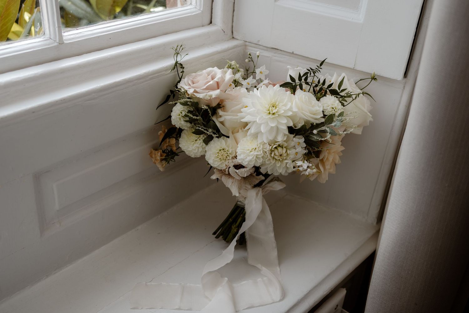 A delicate white and blush wedding bouquet with ribbons sits on a white windowsill in natural light.