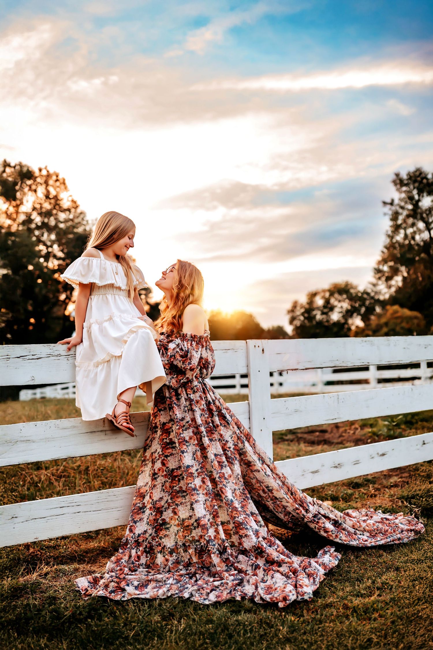 A heartwarming sunset scene with two people in flowing dresses embracing by a white fence in a grassy field.