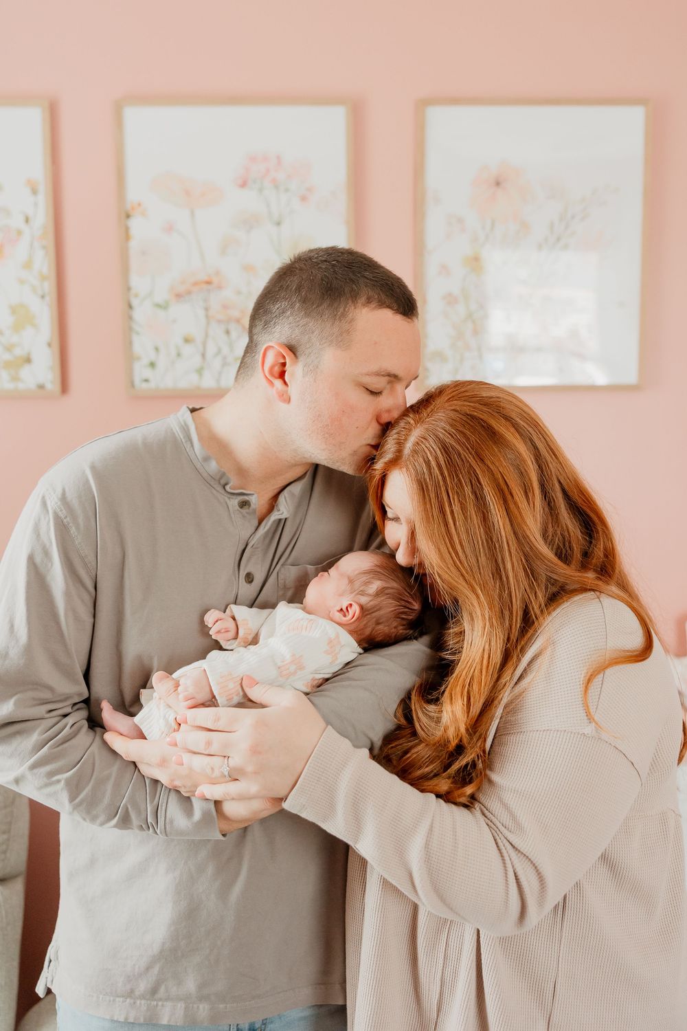 Mom and dad stand together holding their newborn daughter while dad kisses her head.