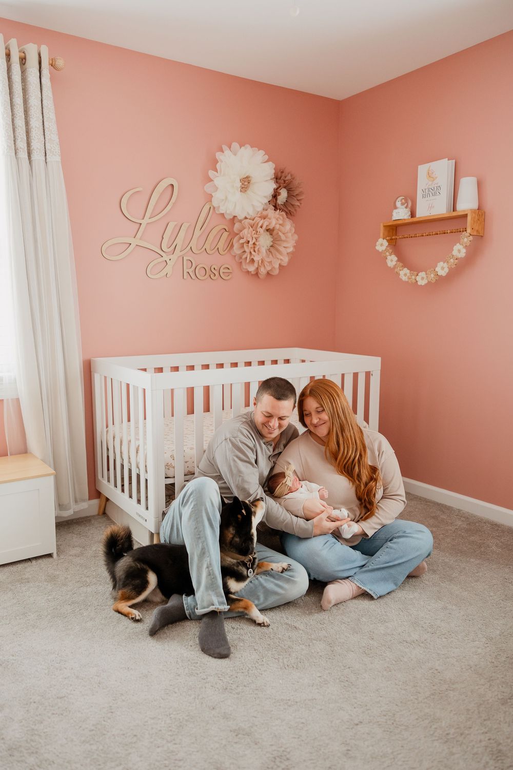 Mom and dad sit on the nursery floor holding their newborn daughter while their dog rests beside them.