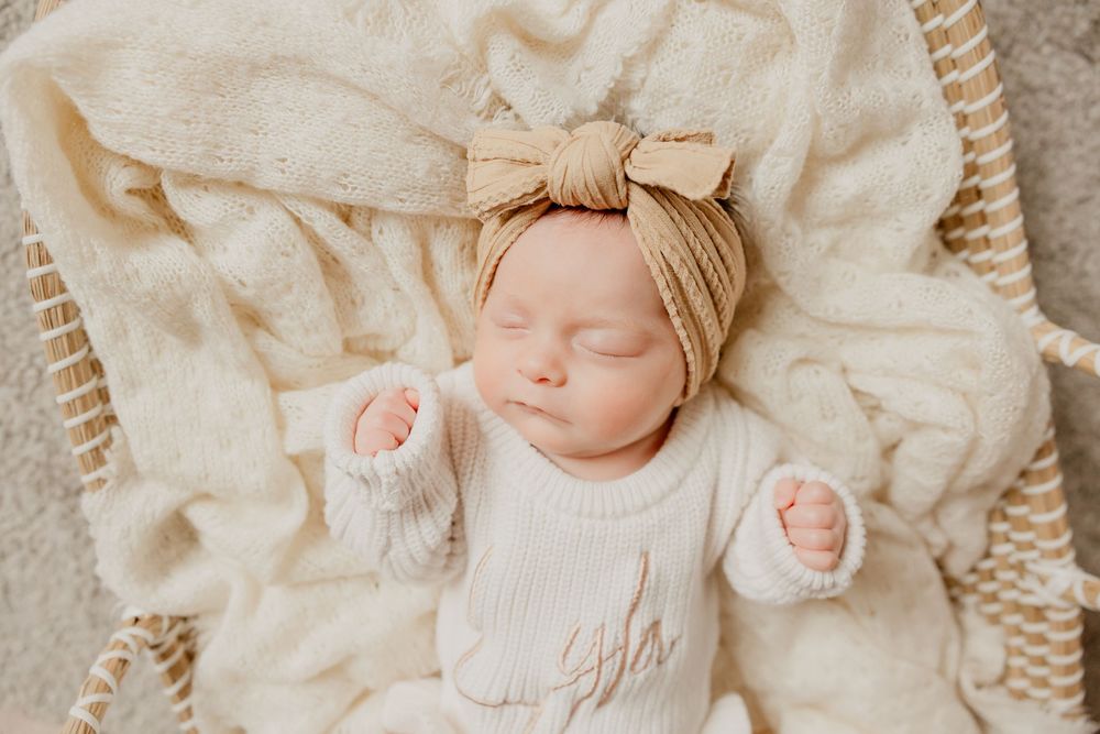Newborn baby girl sleeps on a soft blanket in a basket while wearing a headband.