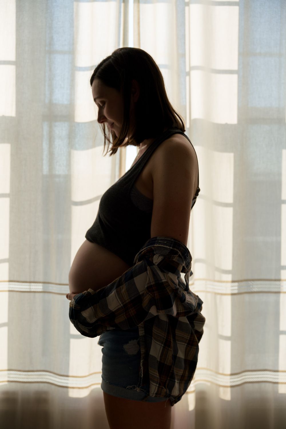 Silhouette of pregnant woman in shorts and plaid shirt standing by window.