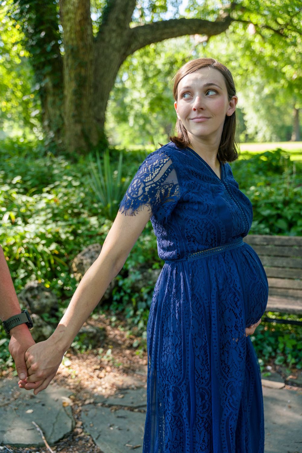 An expectant mother in a blue lace dress poses in a lush garden setting.