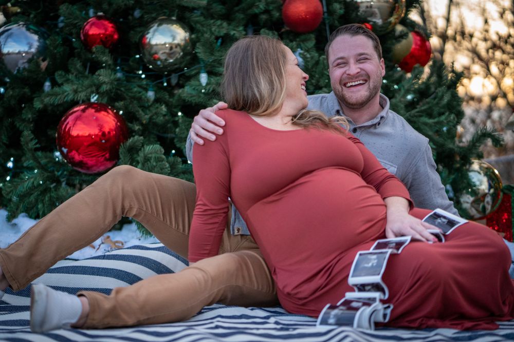 Holiday maternity photoshoot with couple relaxing on blanket by Christmas tree.