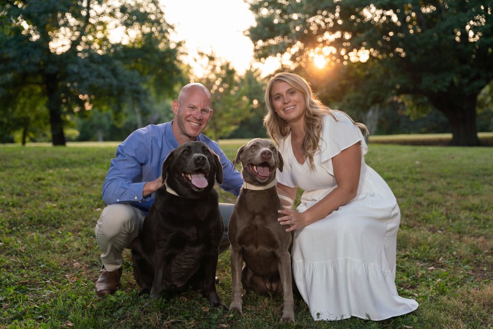 A couple sits with their two chocolate Labrador dogs on grass during sunset in a park setting.