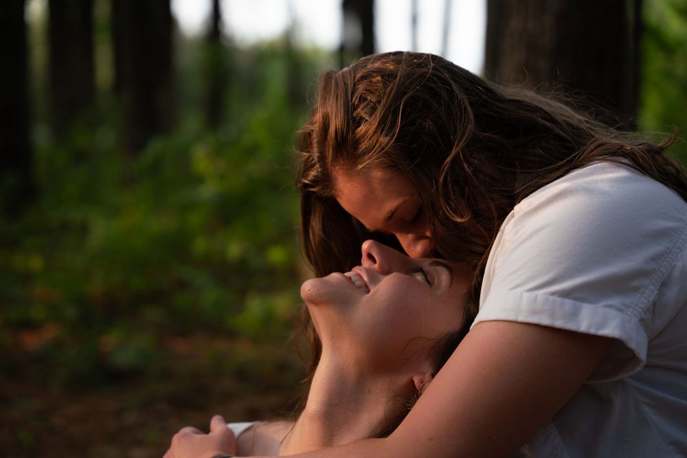 Two people sharing a tender kiss in a sunlit forest with trees and greenery in the background.