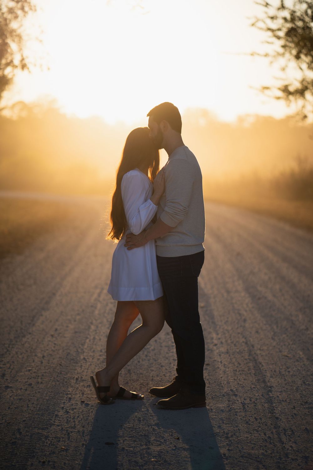 A couple embraces on a dirt road during golden hour sunset creating romantic silhouettes in the warm light.