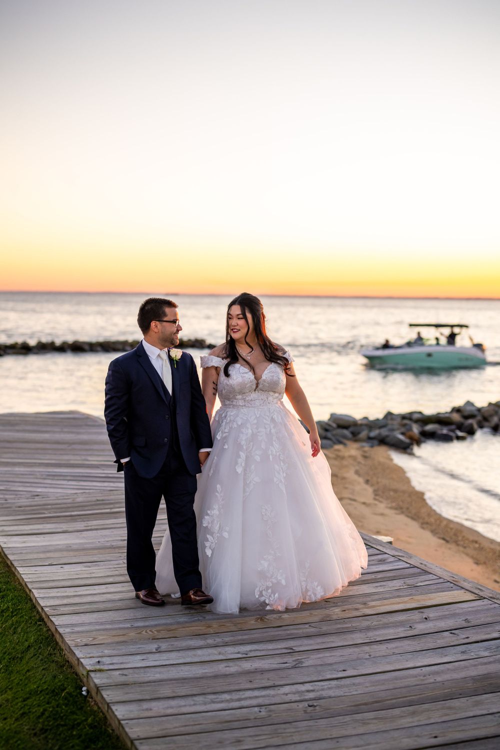 Wedding couple poses on oceanfront boardwalk during sunset with boat in background.