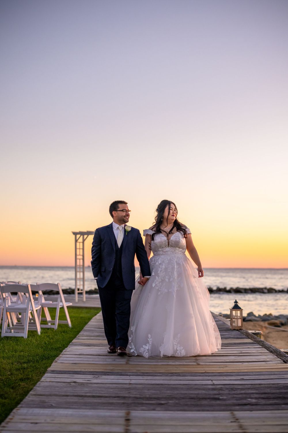 Newlyweds walking along coastal boardwalk at sunset with ceremony setup visible.