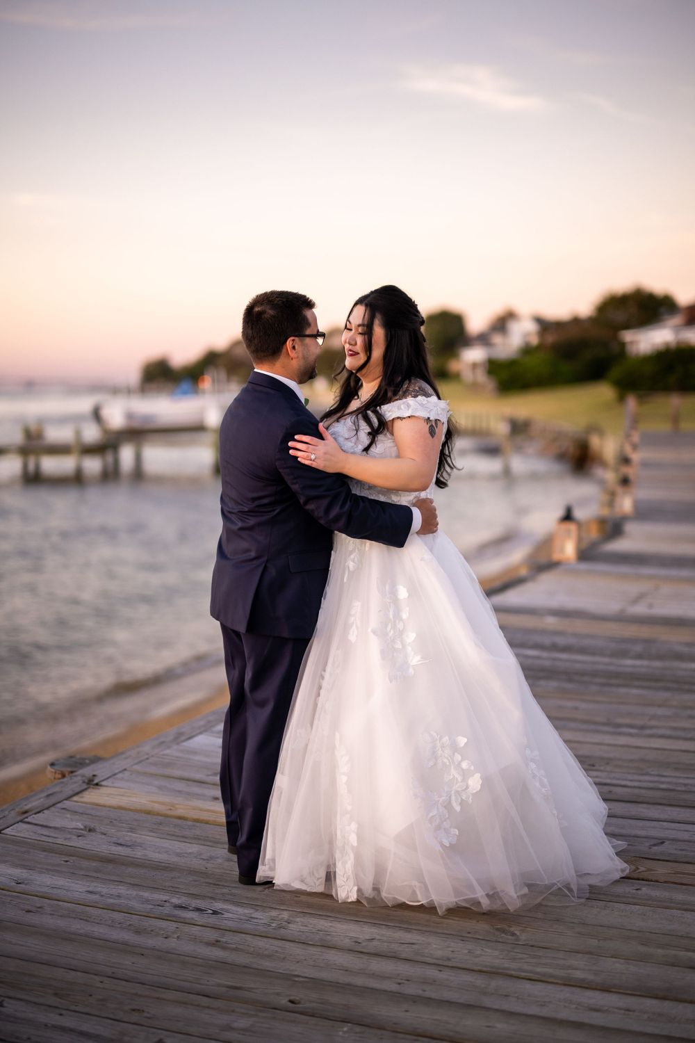 Wedding couple shares intimate moment on wooden boardwalk by the water at dusk.