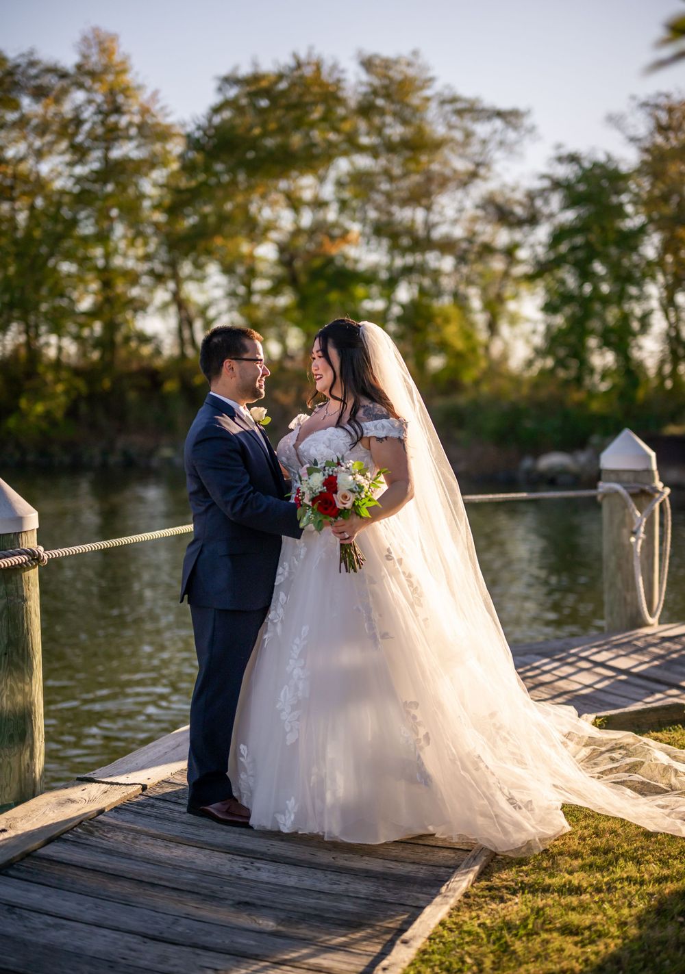 A newlywed couple shares a romantic moment on a wooden dock by a lake at sunset.
