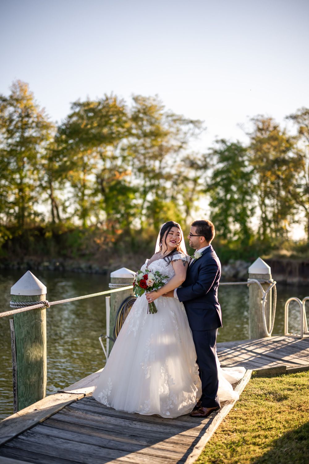 A couple in wedding attire embraces on a lakeside dock surrounded by autumn trees.