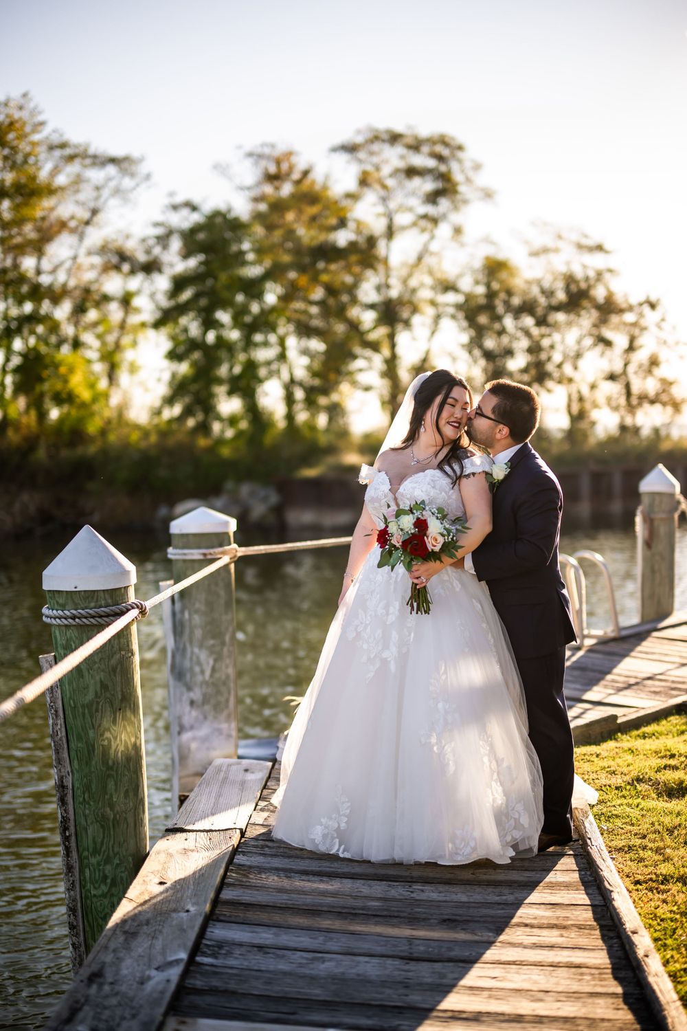 A tender wedding moment captured on a wooden pier with water and trees in the background.