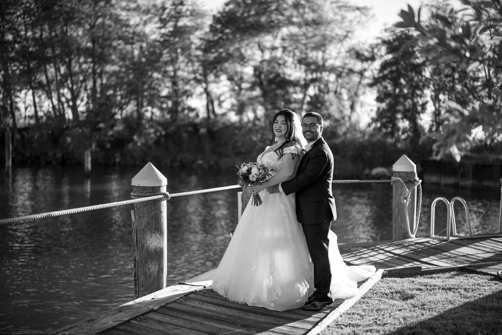 Black and white wedding photo of a couple standing on a wooden dock by the water.