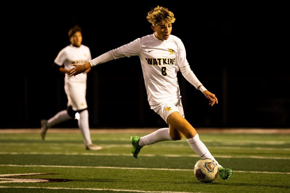 Soccer player in white uniform dribbles ball during nighttime match on illuminated field.