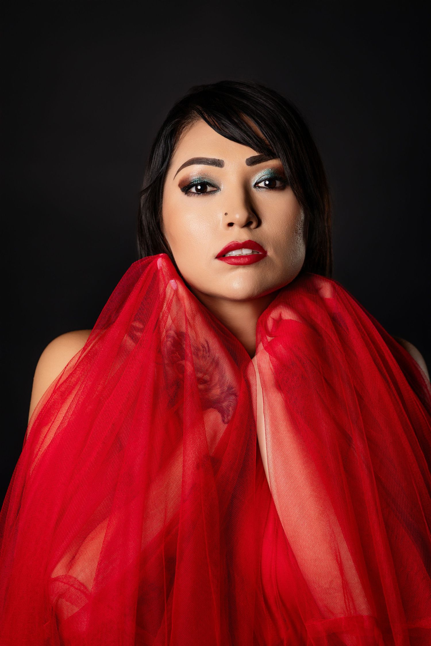 Dramatic portrait with flowing red fabric and bold makeup against black background.