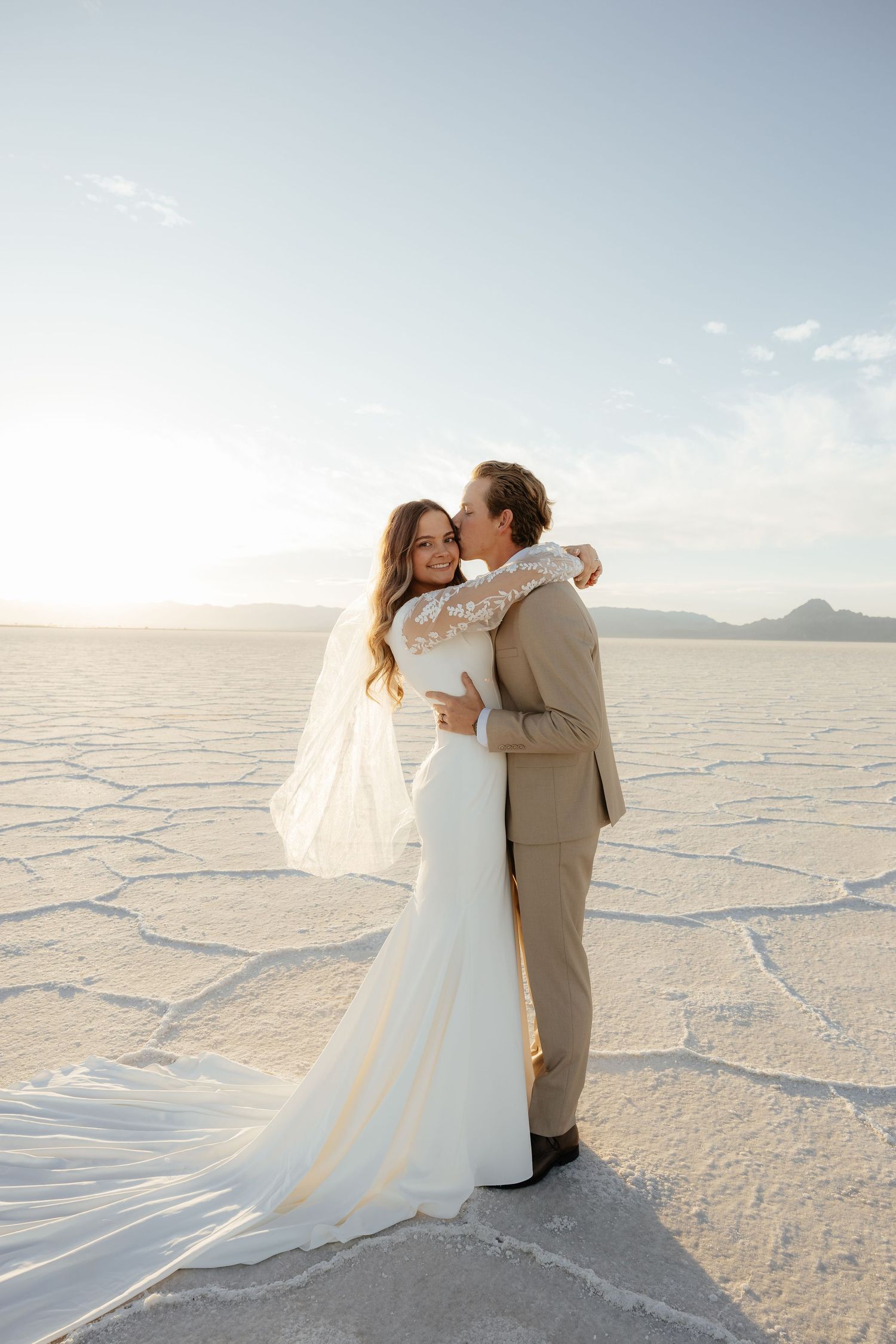 Bride and groom embrace on the Bonneville Salt Flats at sunset, veil flowing in the breeze.