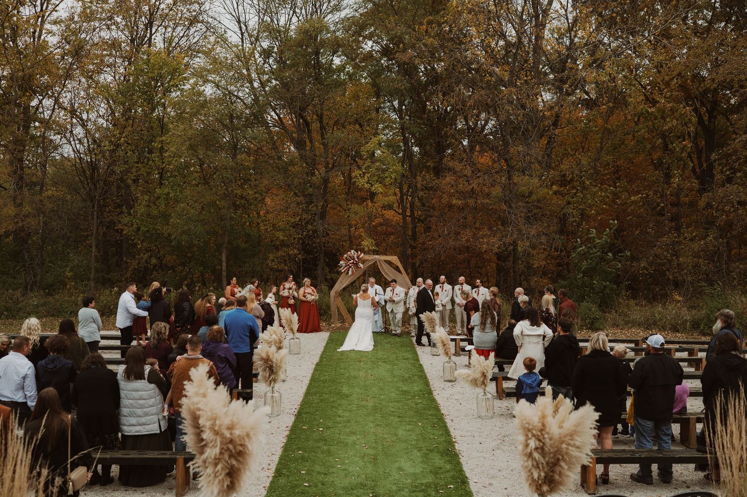 Black and white outdoor wedding ceremony by a fountain with guests seated in white chairs on a lawn.
