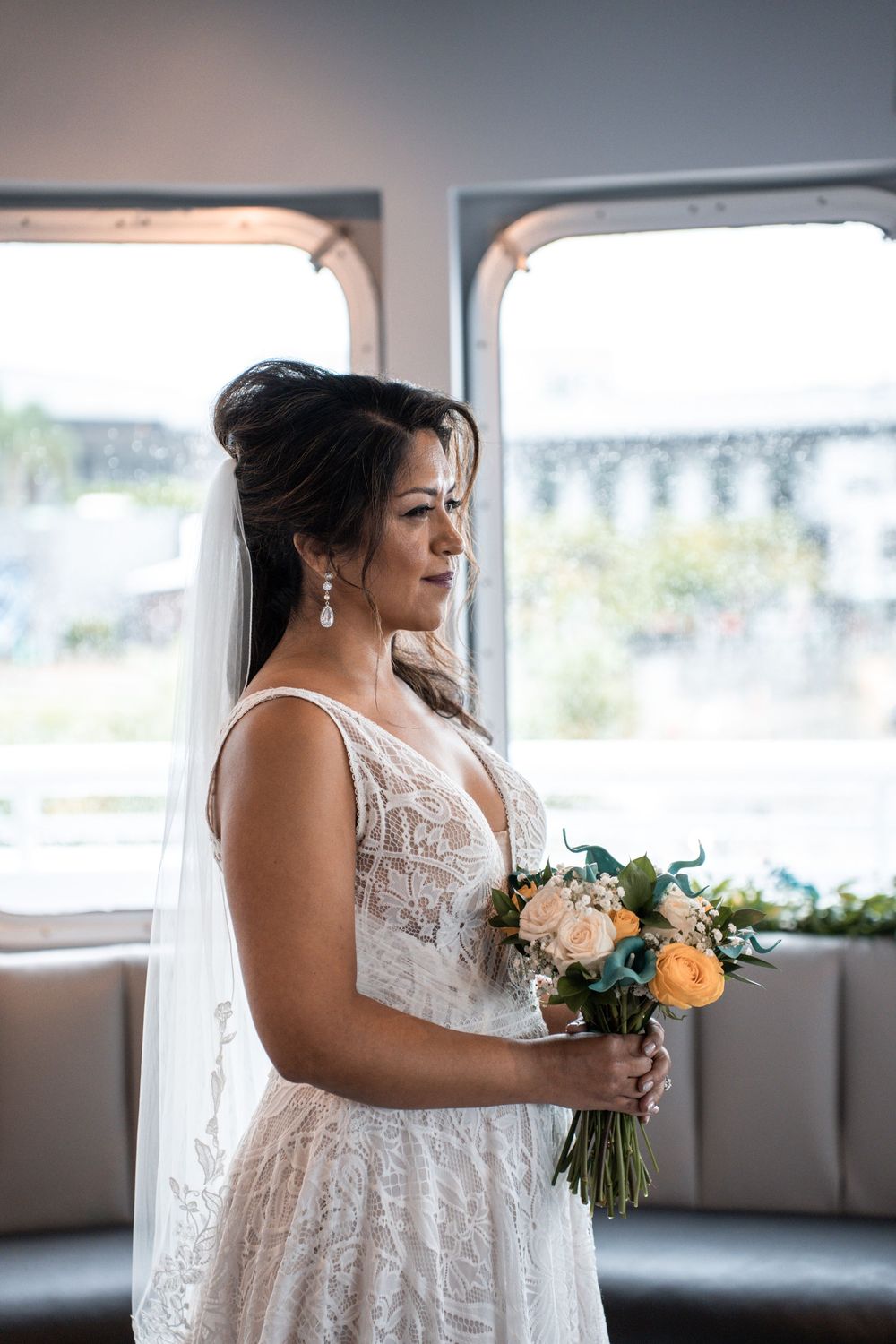 A person in a white lace wedding dress holds a peach and green floral bouquet while standing near a window. Micro wedding on Yacht StarShip