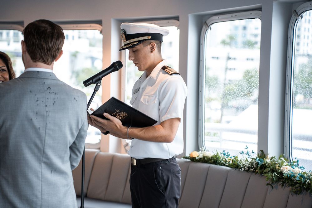 A ship captain in white uniform holding a book during a ceremony on a boat with floral decorations. Micro wedding on Yacht StarShip in Tampa