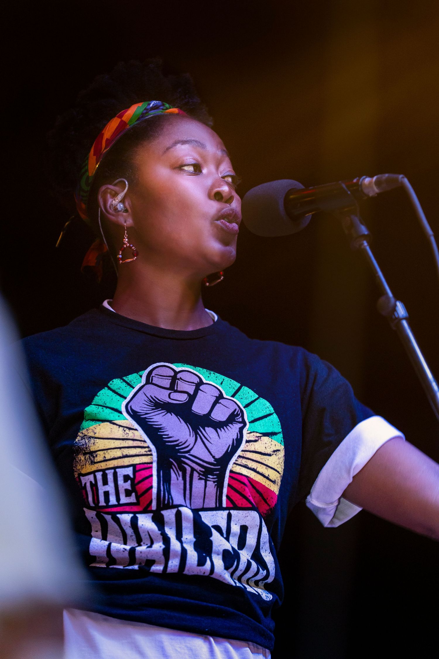 A performer wearing a colorful graphic t-shirt sings passionately on stage in dramatic lighting.