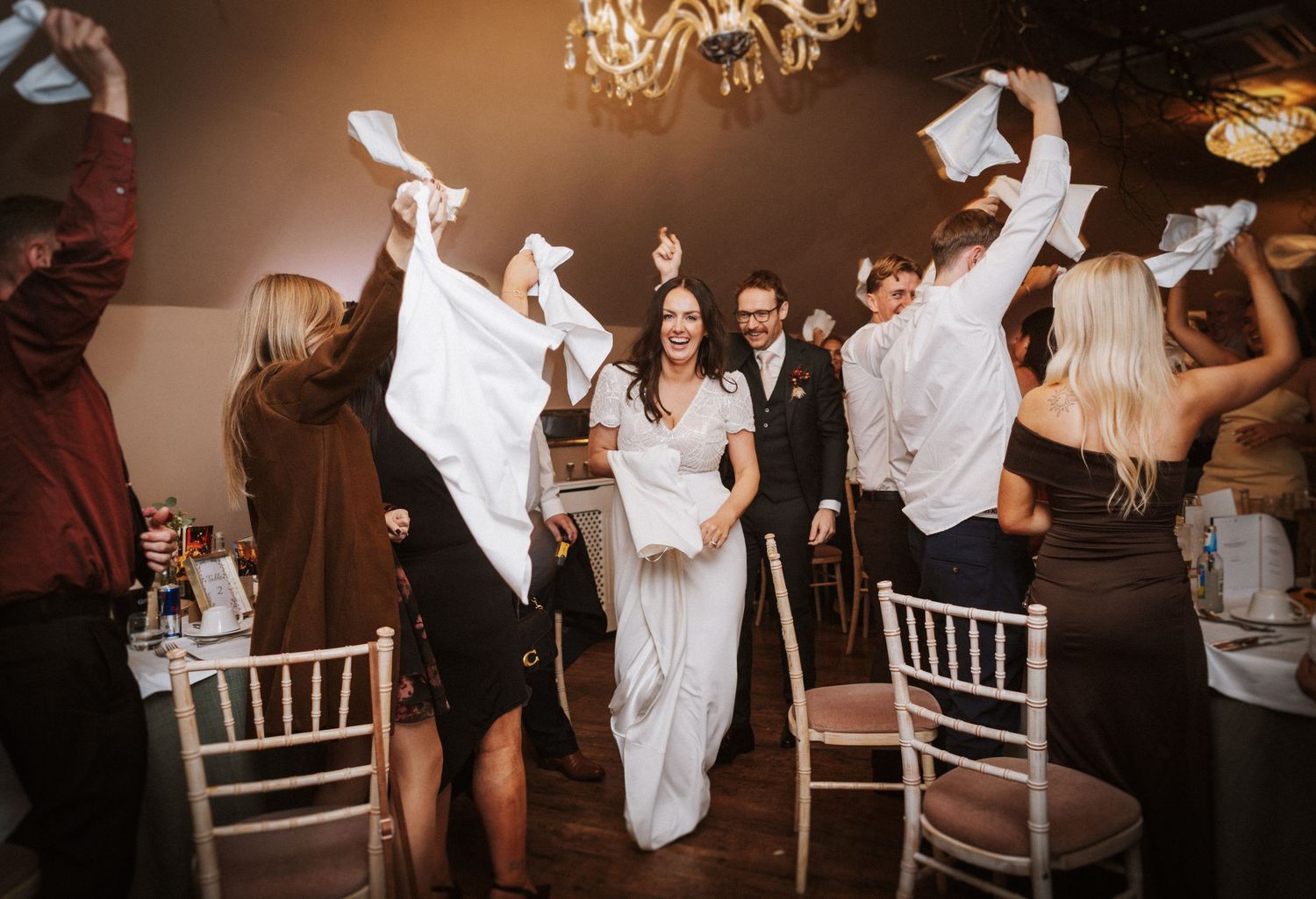 A group dances and celebrates during a lively wedding reception with white chairs and warm lighting.