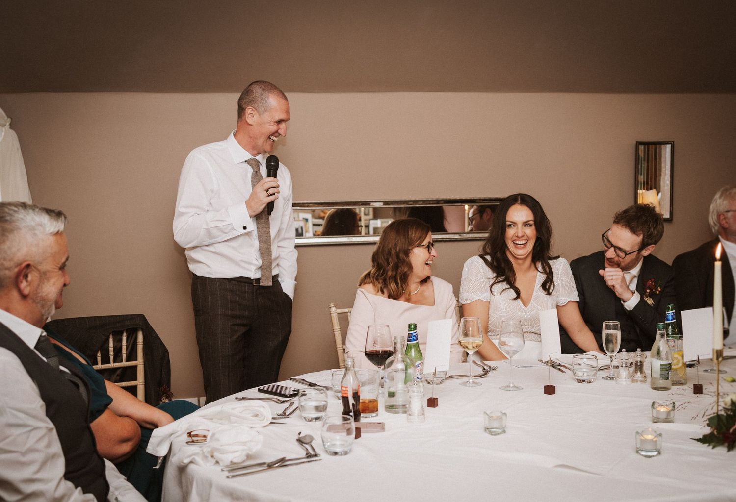 Wedding guests laugh and enjoy themselves during dinner reception at tables decorated with white linens and centerpieces.