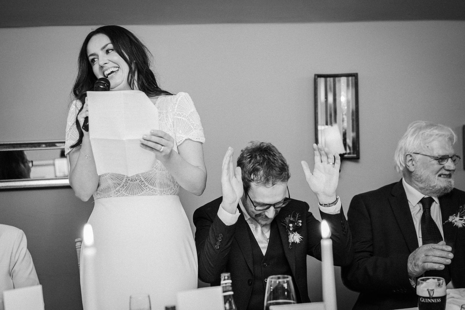 Black and white candid photo of wedding reception guests laughing and raising hands during speeches.