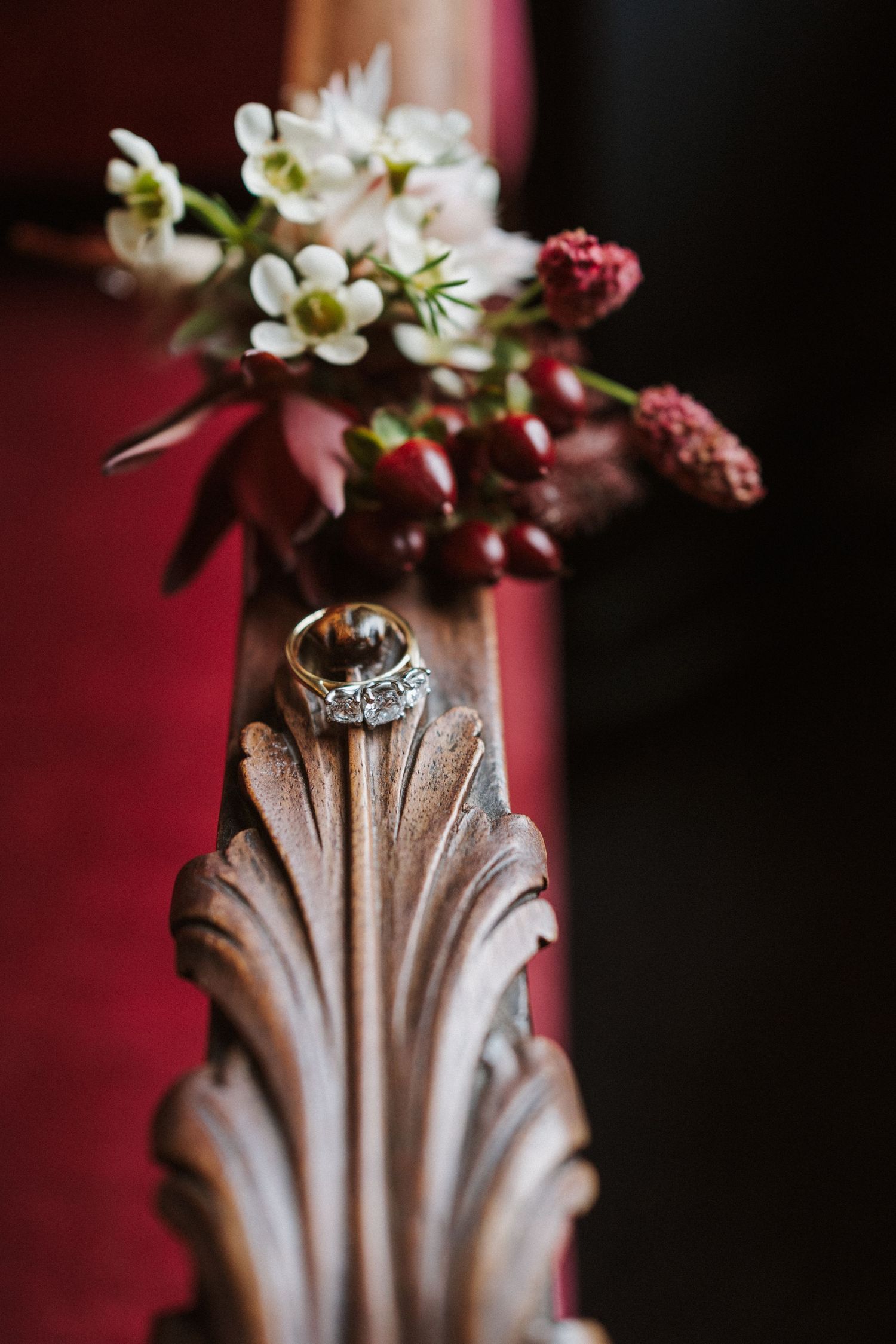 Wedding ring displayed on ornate wooden detail with delicate white flowers and red berries in the background.