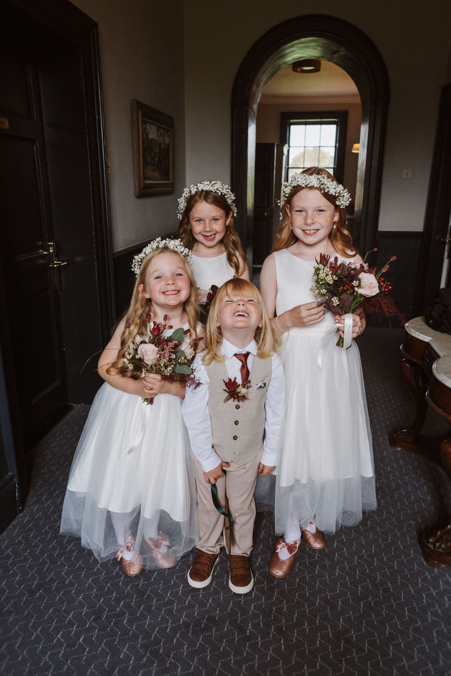 Flower girls and ring bearer in white dresses and tan vest laugh together on grey carpet.