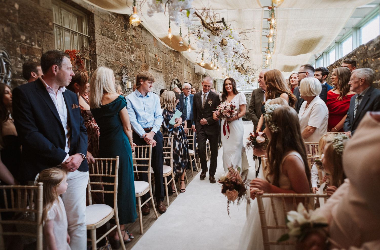 Wedding ceremony guests seated in rows at a rustic stone-walled venue with natural lighting.