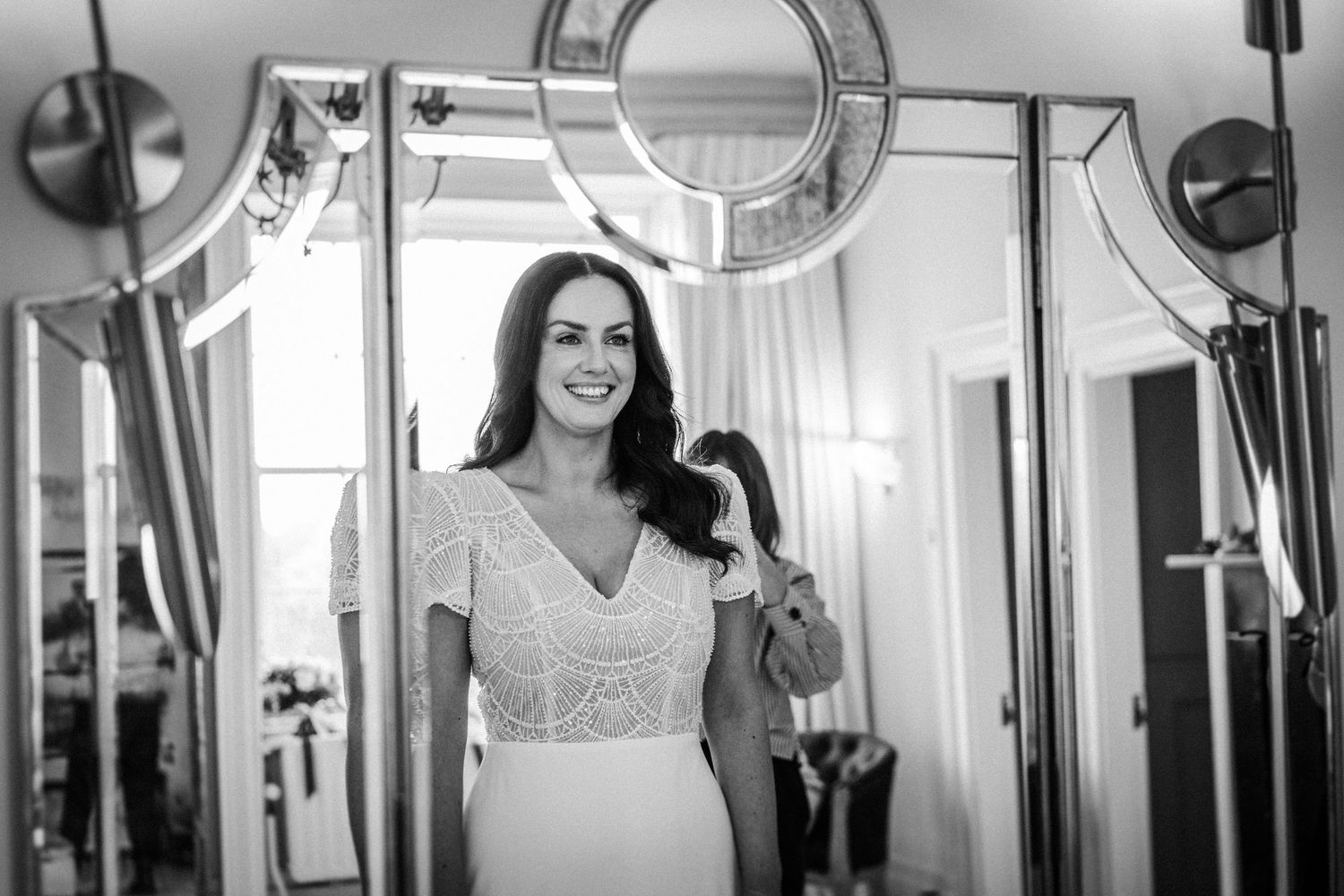 Elegant bride in a white lace dress poses in front of large windows in a bright interior.