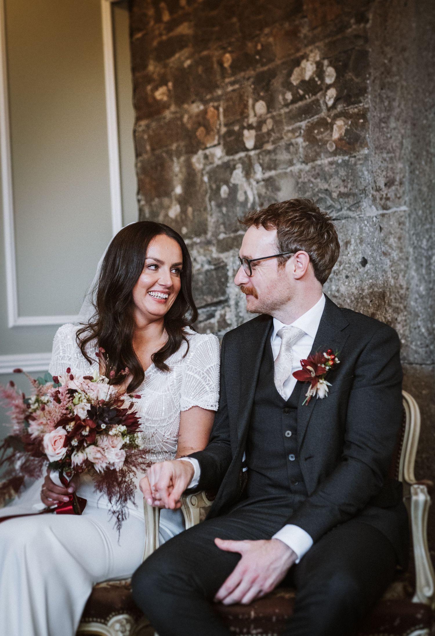 A romantic wedding moment with the couple sharing laughter while seated against a textured wall.