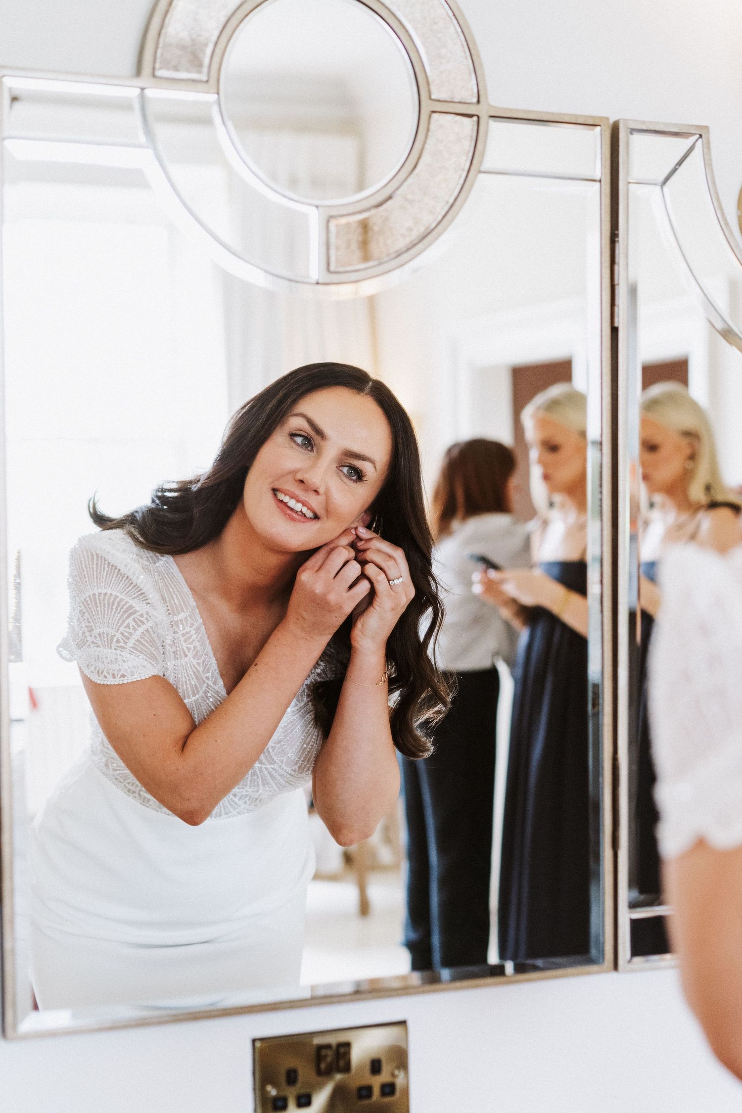 A bride in a white lace dress smiles while getting ready in front of mirrors.