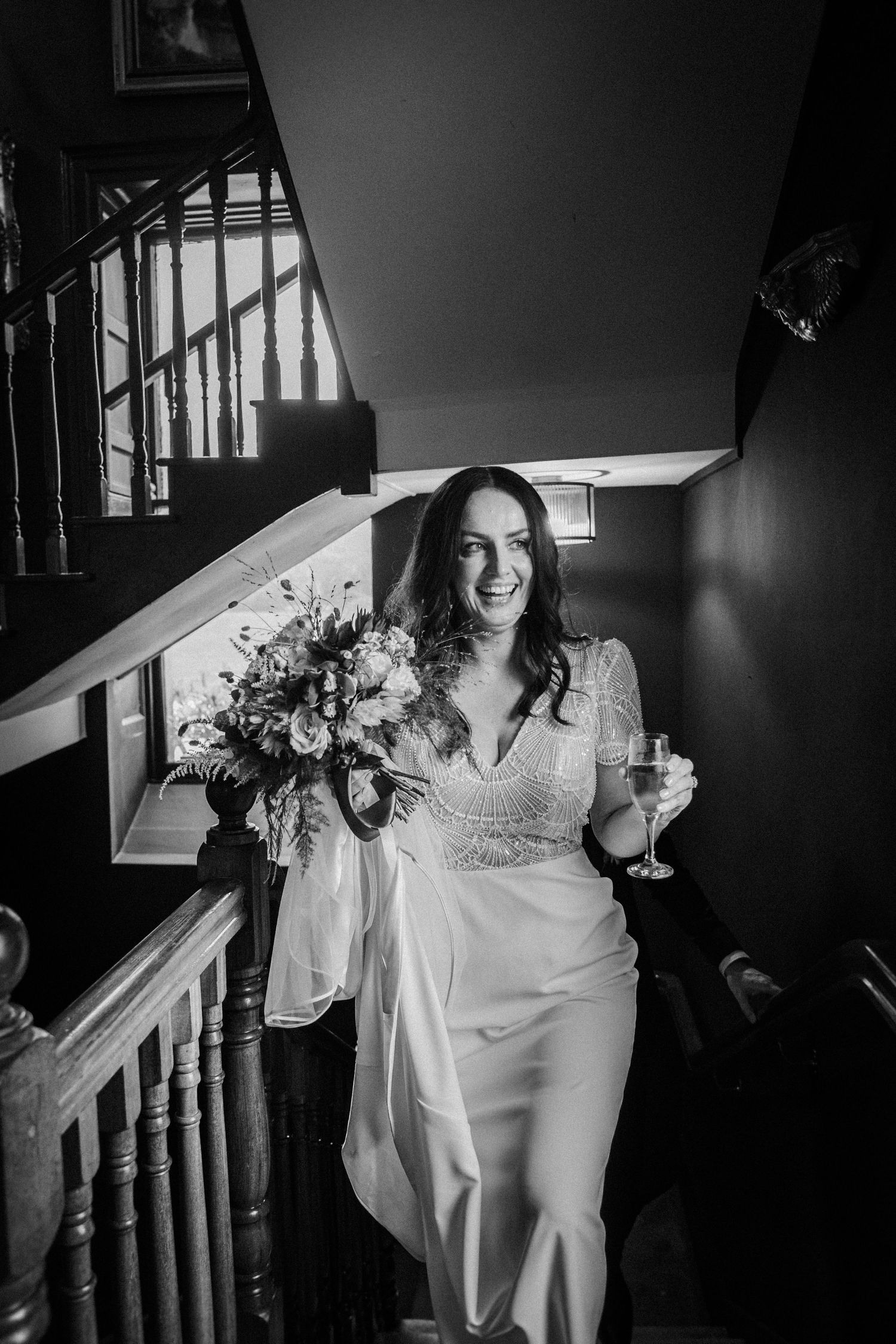 Black and white photo of someone descending stairs in a flowing white dress with flowers.