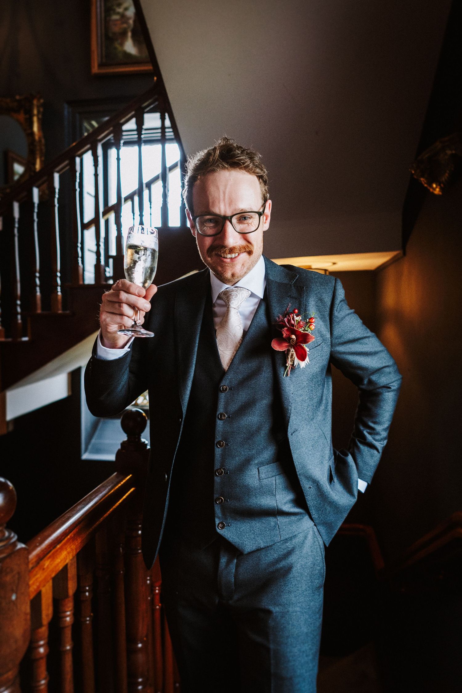 Artistic portrait of groom in grey suit raising glass on wooden staircase in moody lighting.