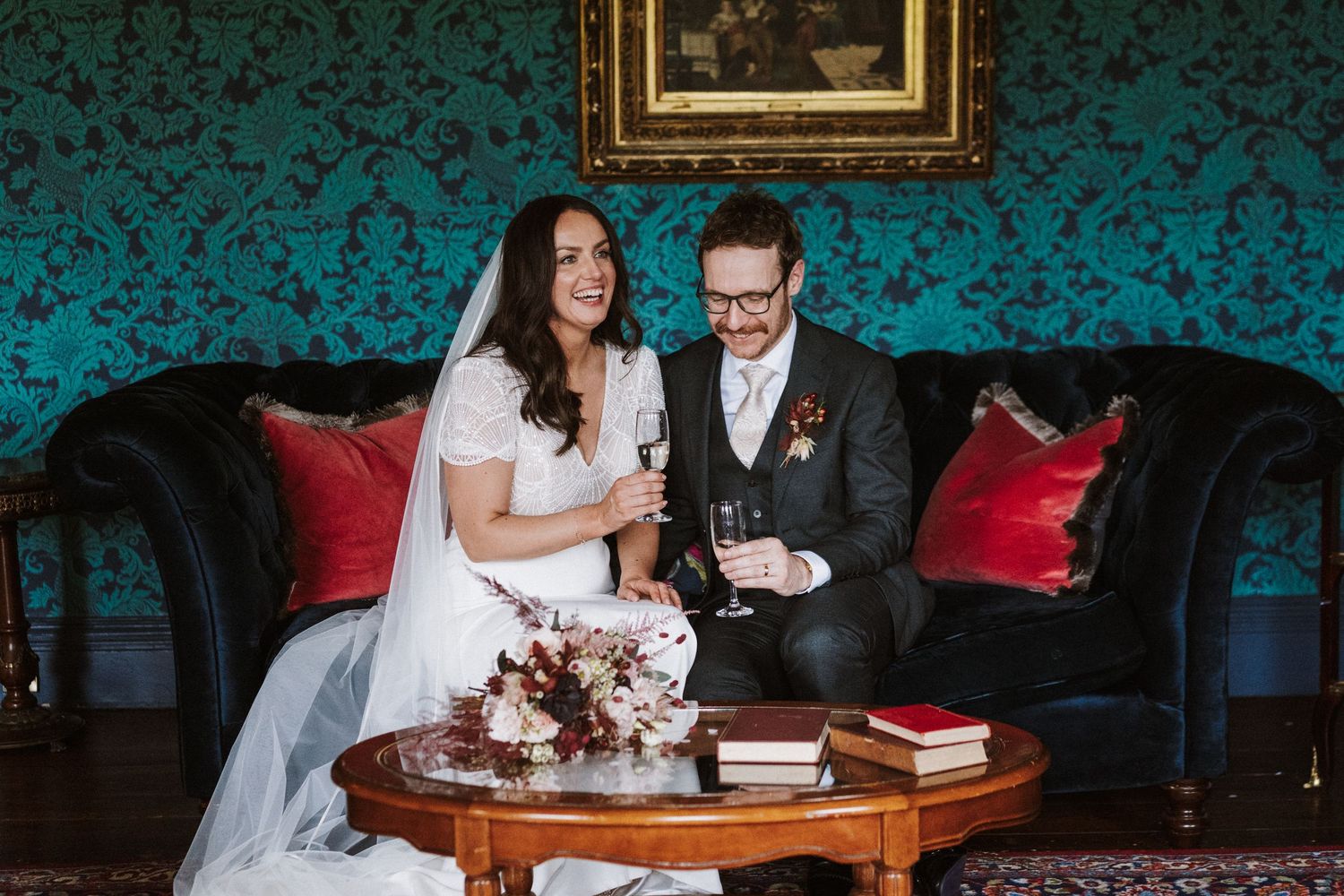 A wedding couple signs documents at an elegant wooden table in a vintage-styled room with teal wallpaper.