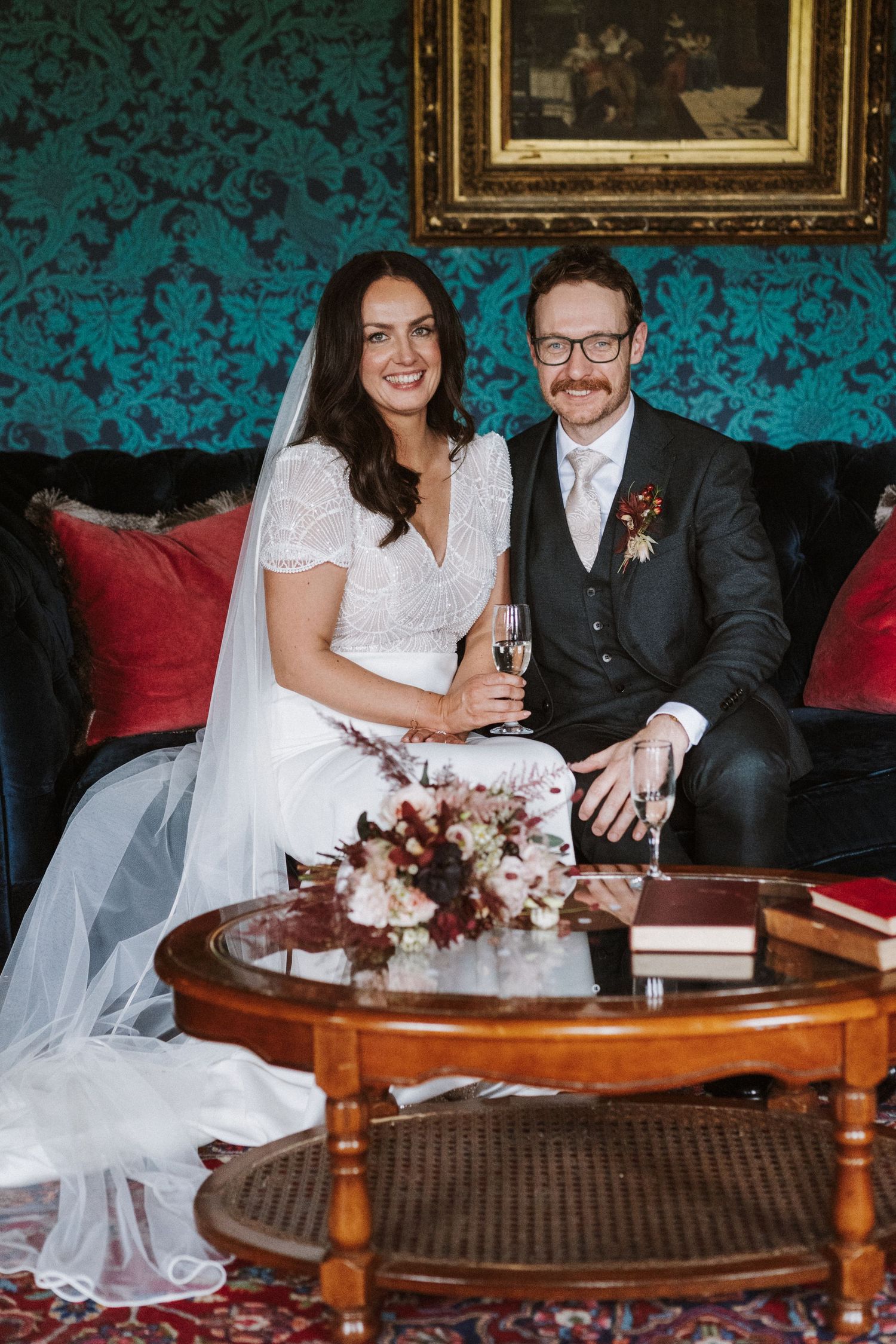 Newlyweds sit together on red furniture against a teal damask wallpapered wall.
