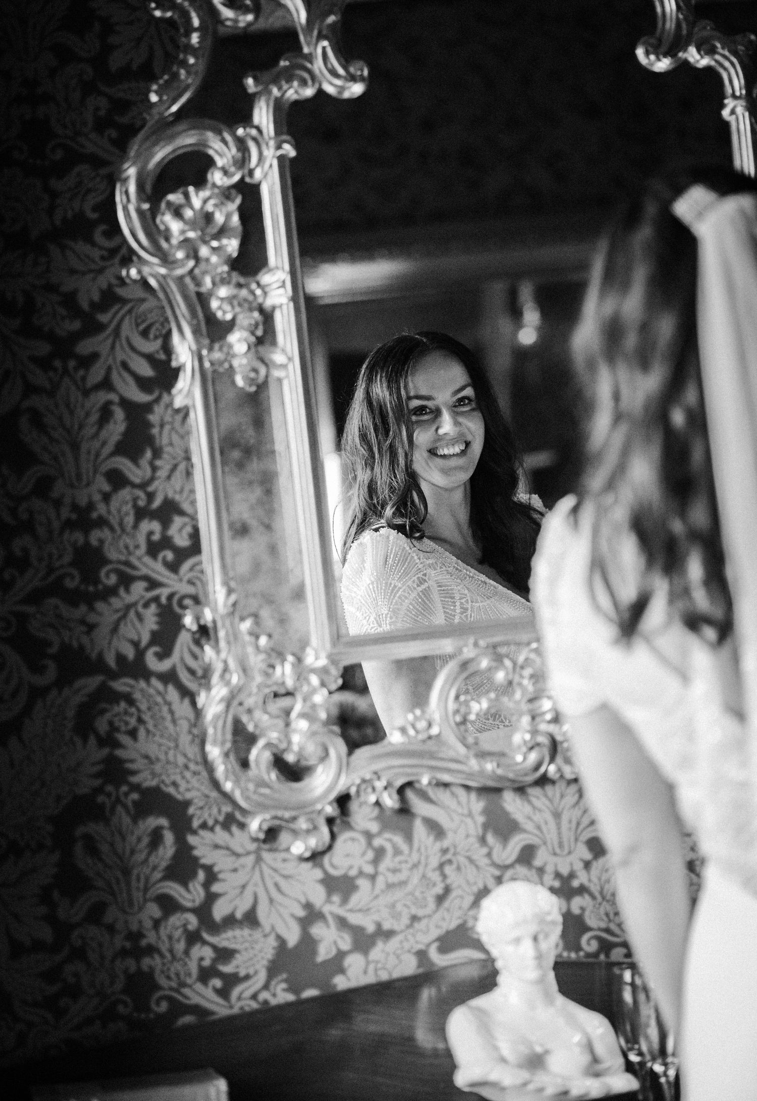 Bride looking at her reflection in an ornate mirror against patterned wallpaper.