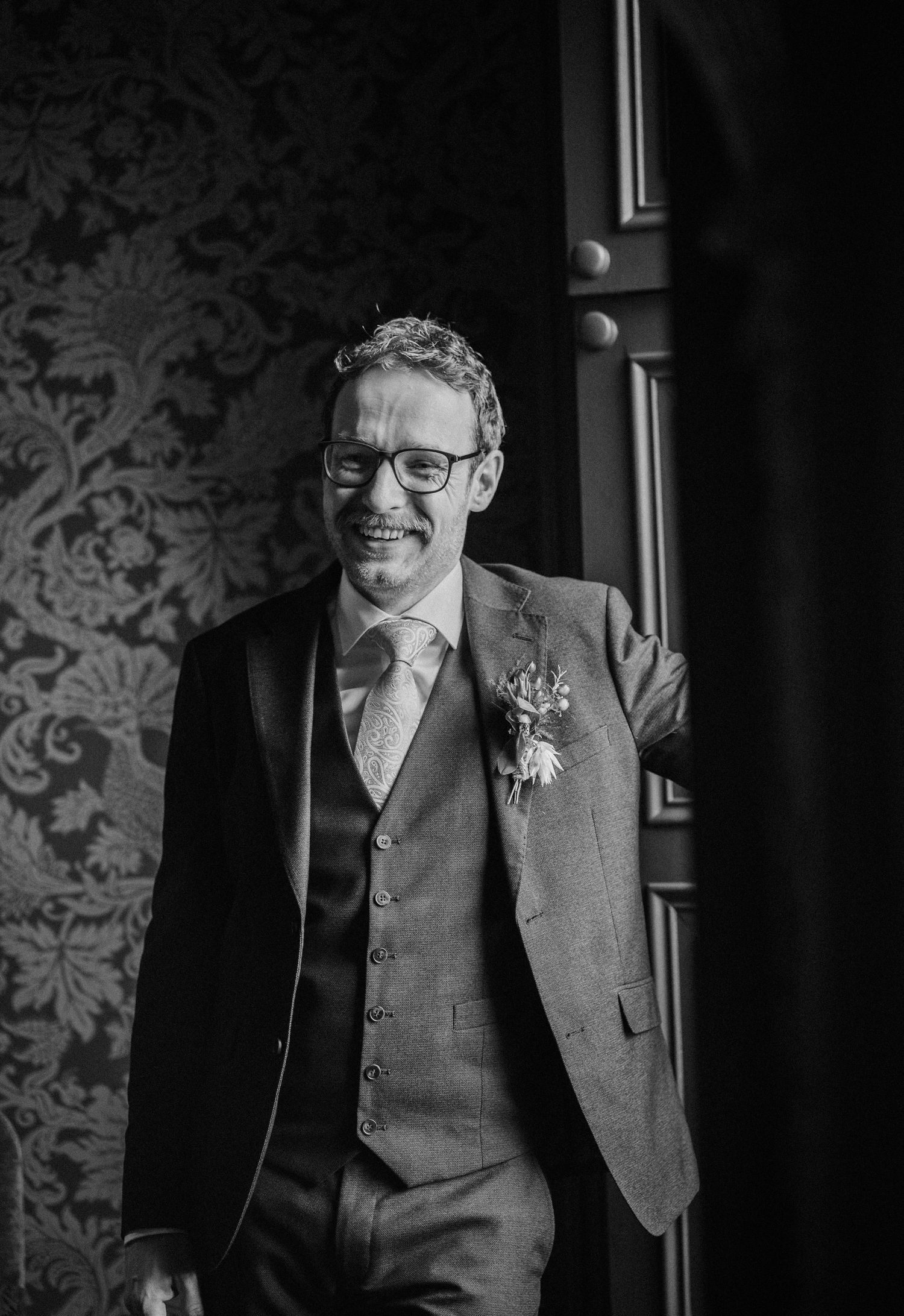 Black and white portrait of a person in formal attire against a patterned wallpaper background.