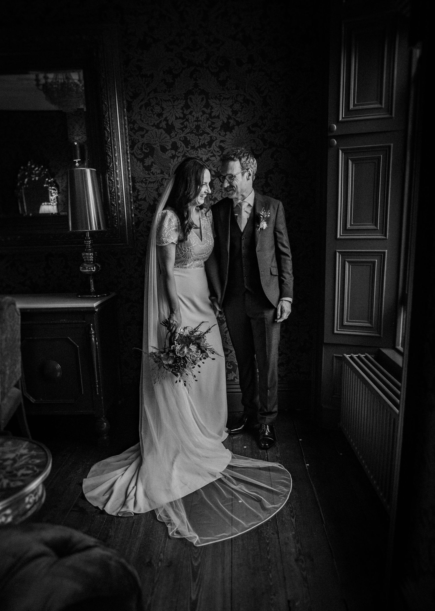 A dramatic black and white wedding photo captures a bride's flowing dress in a dimly lit vintage room.