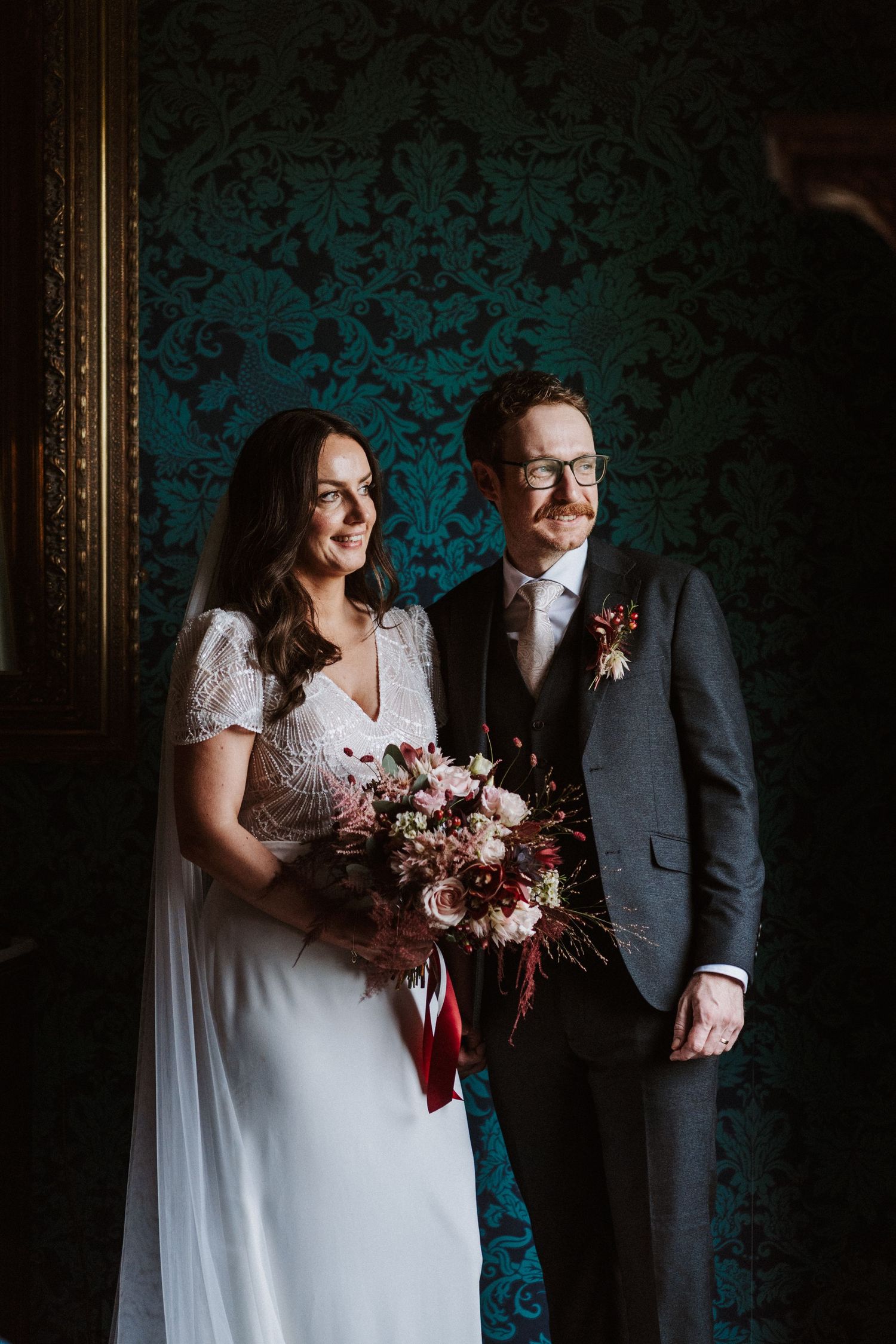 Wedding portraits against teal damask wallpaper with dramatic lighting and floral bouquet.