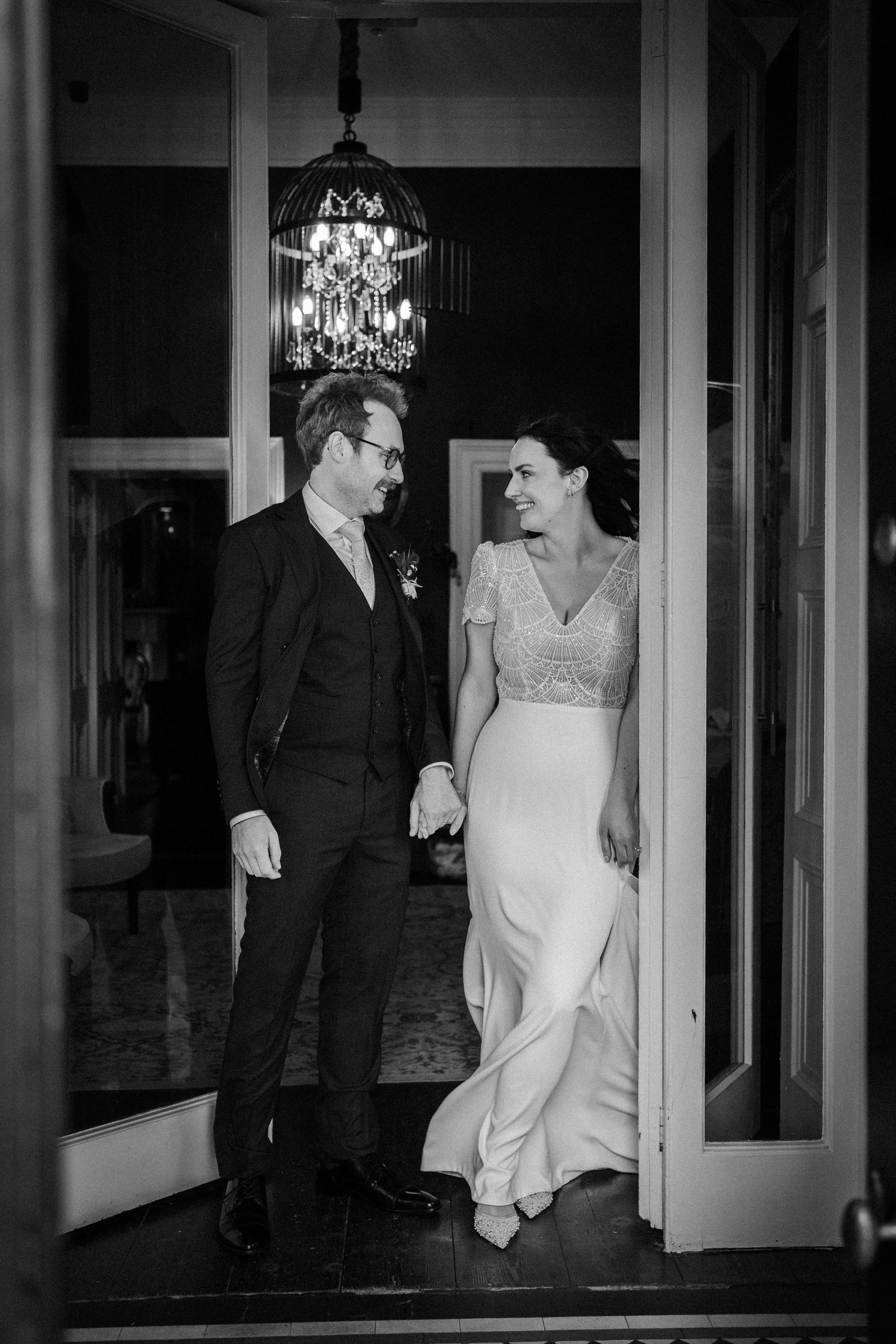 A black and white wedding photo sequence showing a couple walking through elegant doorways under chandeliers.