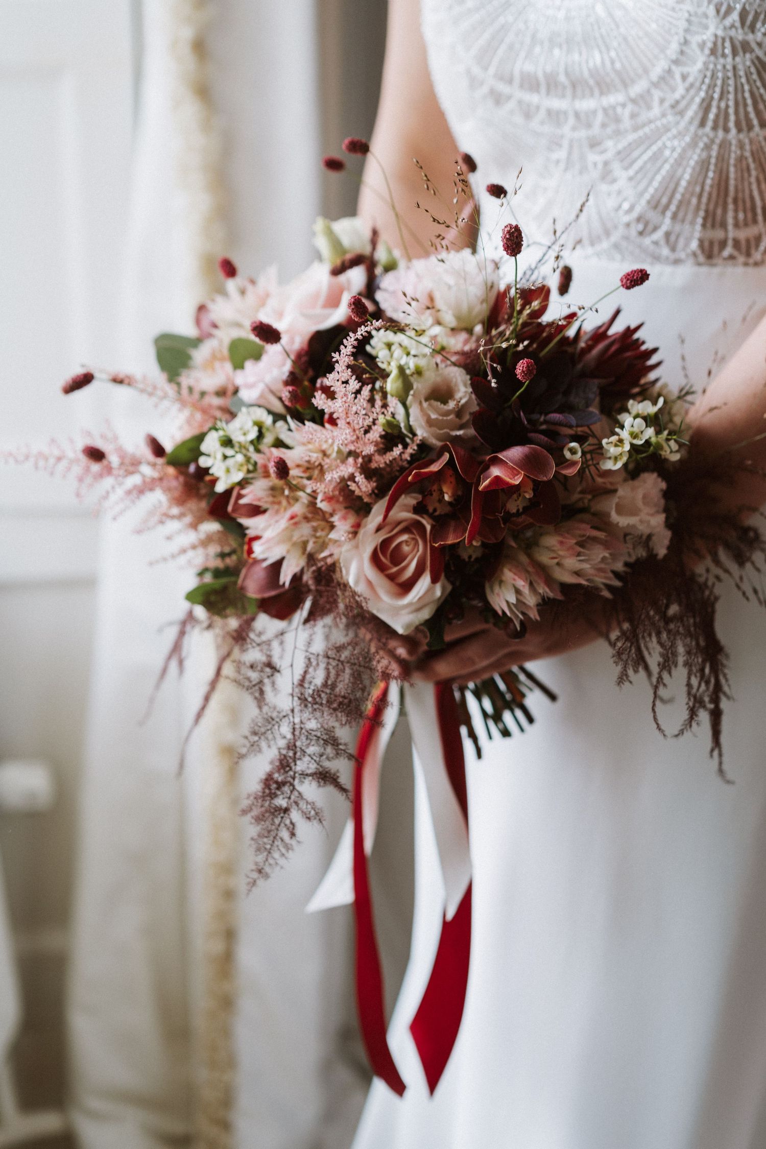 Elegant bridal bouquet featuring blush roses and burgundy ribbons against white lace.