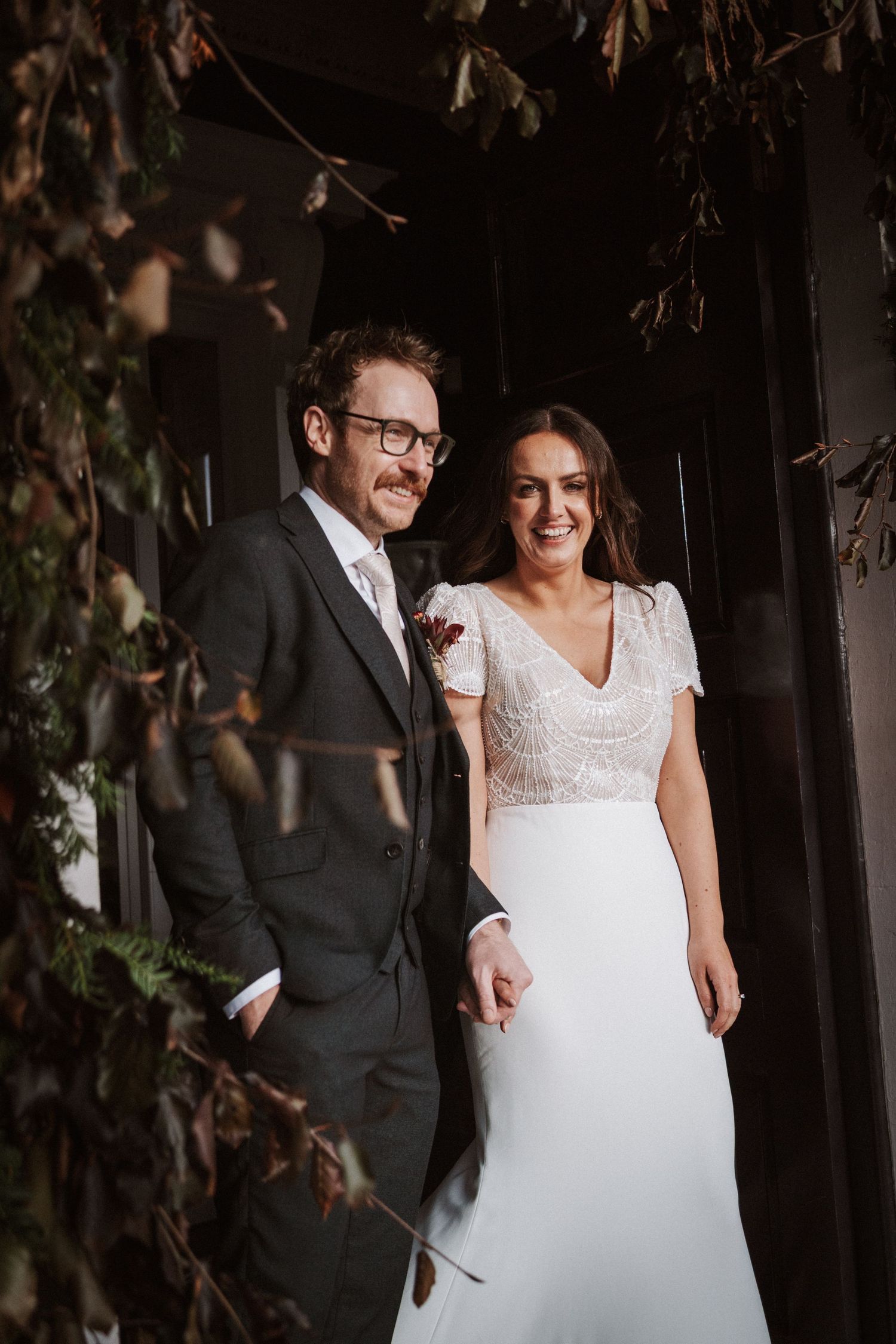 A romantic wedding couple poses together under autumn leaves in a moody outdoor setting.