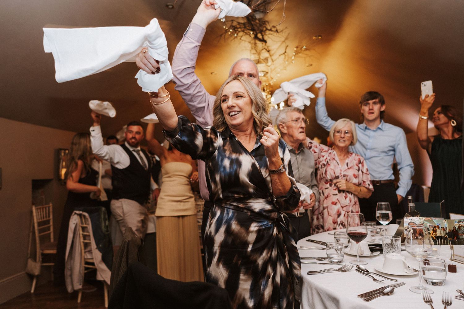 Wedding guests celebrate joyfully during a reception dinner at an elegant indoor venue with warm lighting.