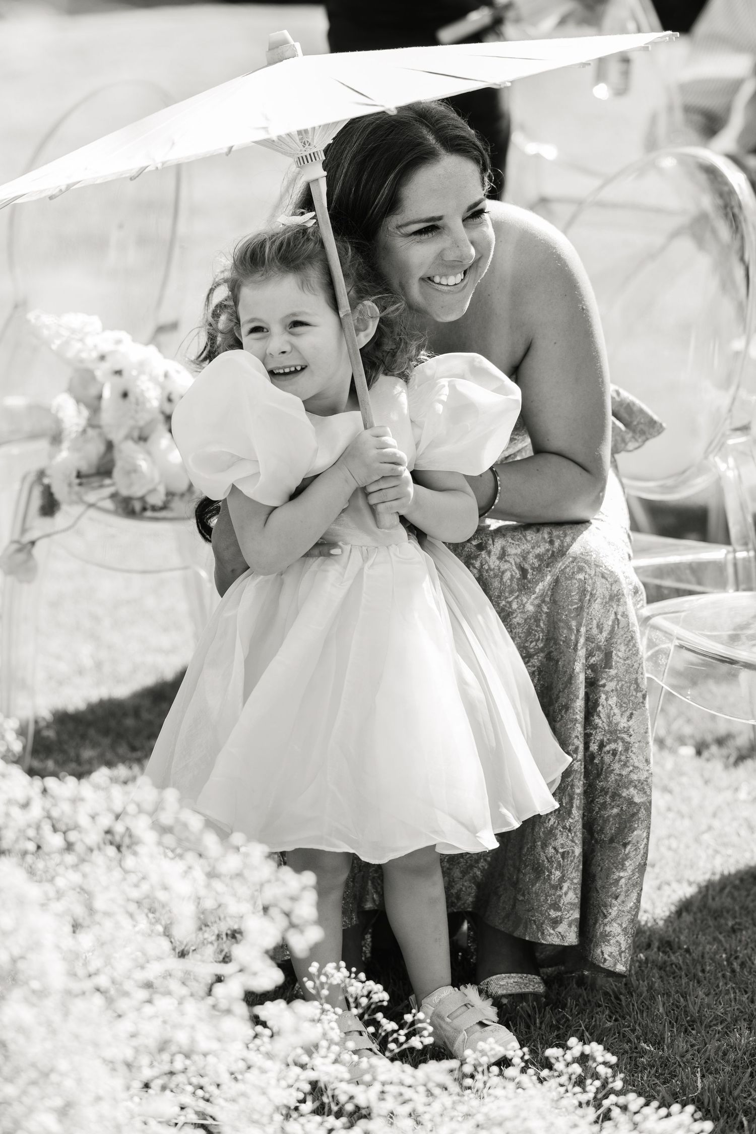 A young child in a white dress holds a parasol while smiling at an outdoor event.